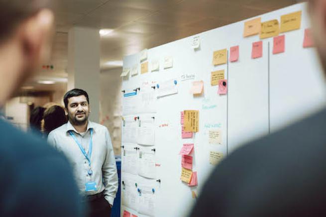 Man stands next to board with post it notes with people looking at him