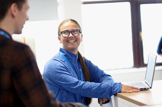 Woman sits at computer in an office talking to a man 